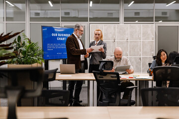 Diverse business colleagues, including a White senior man and an adult Caucasian woman, review documents and presentations around a wooden table during a corporate team meeting in a modern office.