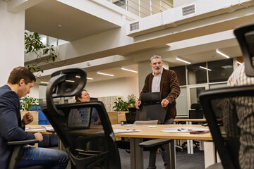 A gray-haired man in his early 50s in a brown jacket is having a meeting with a dark-haired Asian 30-year-old woman and two White adult colleagues in a blue coat and plaid shirt in the office.