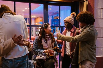 Fototapeta premium A group of multiethnic friends enjoys a cozy evening outside a cafe. Laughter, warm drinks, and lively conversation fill the air as they connect under the city lights. One holds a small dachshund.