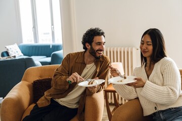 Young man with curly hair and a beard and a young Asian woman sitting in armchairs, eating cake in...
