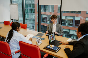 A young Caucasian man in a gray suit sits at a table during a business meeting with two diverse colleagues in the office. They discuss a strategy using documents and a laptop with data visualization.