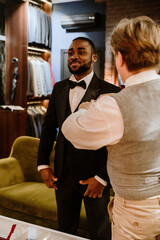 A Black groom in a black tuxedo and with a bow tie stands confidently while a tailor in a vest adjusts his suit. The wedding boutique features elegant suits hanging on racks and a stylish interior.