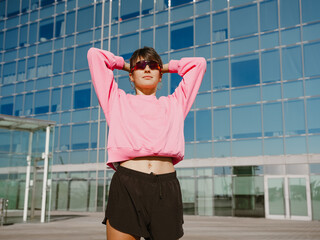 A young female athlete in a pink sweatshirt and black shorts stretches outdoors in front of a modern glass building. The urban backdrop highlights her confidence, energy, and active lifestyle.
