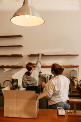 Two white-skinned women, cafe owners, arrange plates and glasses on shelves as they prepare the...
