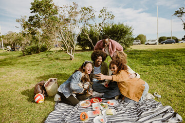 A group of young multiethnic friends enjoys a picnic in the park. They sit on a blanket with fruits, laughing and taking selfies with a smartphone. A small dog sits on one friend's lap.
