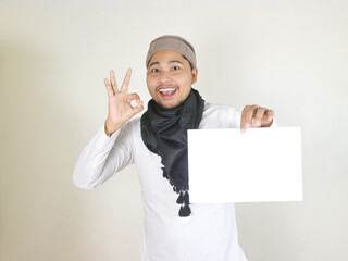 handsome asian muslim man wearing black turban or shemagh holding blank white paper and giving okay...