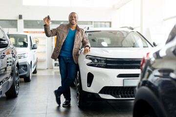In a modern car dealership, a cheerful young African American man stands next to his new white...