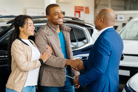 A young African American couple is finalizing a vehicle purchase at a car dealership. They are happily shaking hands with the salesman while reviewing the contract together in the showroom.