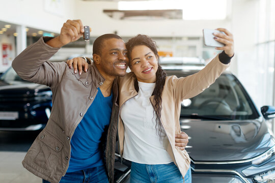A happy African American couple stands in a car dealership, holding keys and taking a selfie together to celebrate their new vehicle purchase. The atmosphere is joyful and festive.