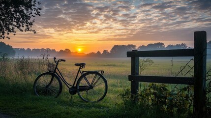 Fototapeta premium A scenic countryside view with a classic bicycle parked beside a wooden fence, overlooking a vast meadow at sunset.