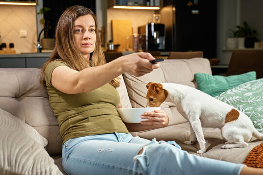 Woman relaxing on sofa with remote control while dog steals food. Pet dog eat food from bowl while owner focuses on TV. Home life and pet behavior concept - Powered by Adobe