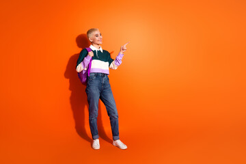 Young stylish woman with a backpack pointing to the side against a bright orange background