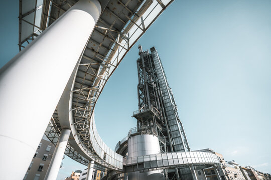 Low-angle view of modern industrial architecture featuring steel beams, curved elevated walkway, futuristic metallic structures, and buildings against clear blue sky background; wide-angle shot
