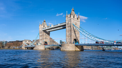 Obraz premium Tower Bridge stands majestically against a clear blue sky, showcasing its iconic architecture. People can be seen enjoying the waterfront, while boats navigate the Thames River.