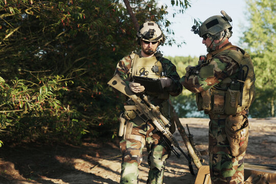 The military unit, dressed in camouflage with rifles slung over their shoulders, reviews mission details on a tablet while coordinating with the army base for the upcoming field exercise.
