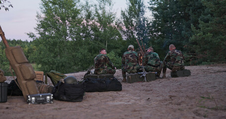 A group of hunters in full camouflage sits around a campfire, grilling sausages and exchanging...