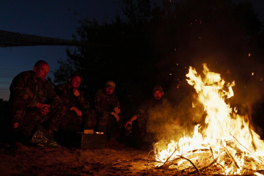 A large bonfire flame, behind it military men in camouflage uniforms are sitting, talking and warming themselves at night by the hearth
