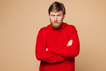 Fototapeta premium Young frowning mad sad Caucasian man he wearing red hoodie casual clothes hold hands crossed folded looking camera isolated on pastel plain light beige background studio portrait. Lifestyle concept.