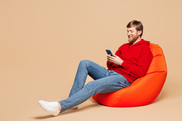 Full body young Caucasian man he wear red hoodie casual clothes sit in bag chair hold in hand use...