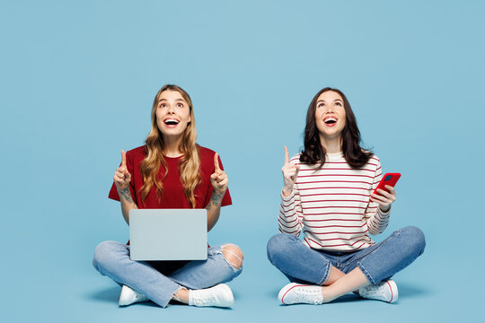Full body young fun couple two IT friends women wearing red casual clothes together sit hold work on laptop pc computer use mobile cell phone point overhead isolated on pastel plain blue background.