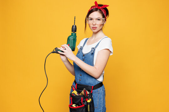 Young employee laborer handyman woman wears overalls belt hold use electric power drill isolated on plain yellow background. Instruments accessories for renovation apartment room. Repair home concept.
