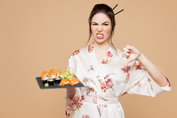 Young displeased sad woman wear kimono clothes hold eat raw fresh sushi roll served on black plate...