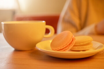 A warm cup of coffee sits beside a plate of colorful macarons on a wooden table in a cozy cafe. The soft afternoon light creates a relaxed atmosphere perfect for indulgence and conversation