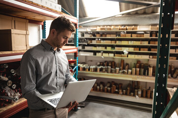 Engineer using laptop in hydraulics factory, inspecting inventory on shelves