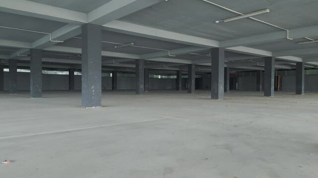 A drone captures an empty underground parking garage with concrete pillars and open sides. The industrial space is illuminated by natural light, creating a modern yet abandoned atmosphere