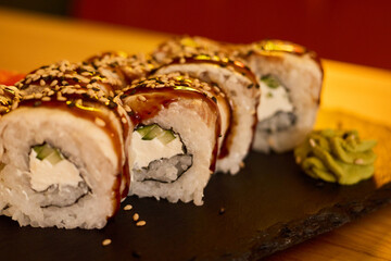 Exquisite sushi rolls are presented on a black slate plate, beautifully drizzled with sauce and adorned with sesame seeds. Vibrant wasabi complements this gourmet dish