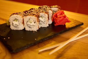 Delicious sushi rolls are arranged neatly on a black slate platter, accompanied by pickled ginger and chopsticks, in a modern restaurant setting during dinner time