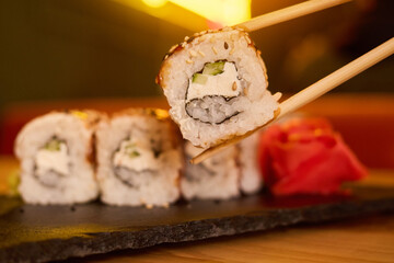 Fresh sushi roll featuring rice, seaweed, cucumber, and cream cheese is enjoyed with chopsticks. The cozy atmosphere is enhanced by soft background lighting