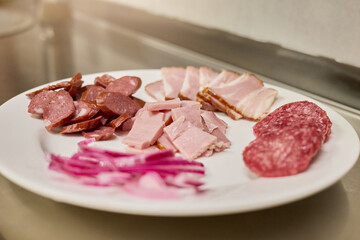 A variety of cold cuts including salami, ham, and bacon are arranged neatly on a white plate. Freshly sliced pickled onions add a pop of color, enhancing the appetizing display