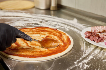 A cook wearing gloves spreads tomato sauce evenly over fresh pizza dough on a stainless steel countertop. Sliced meats are arranged on a nearby plate. Flour dust covers the work surface