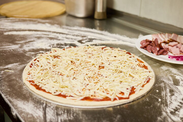 A pizza base topped with rich tomato sauce and generously sprinkled with shredded cheese sits on a stainless steel countertop in a lively kitchen