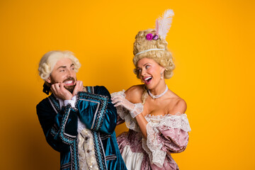 Young couple in historical royal costumes laughing against yellow background showing joyful...