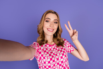 Fototapeta premium Smiling young woman taking selfie with peace gesture wearing pink checkered shirt in front of violet studio background