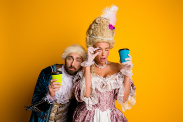 Funny couple in renaissance costumes holding coffee cups against bright yellow background, blending...