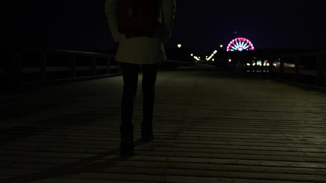 Slow motion grainy evening camera view of woman walking outside. Back view of female legs, stalker pov following his victim from behind. Ferris wheel in the background.