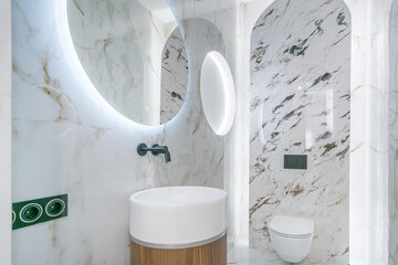 Modern bathroom design, decorated with white marble tiles. Illuminated mirror, stylish accessories.