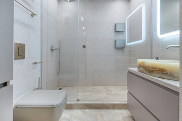 Laconic design of the bathroom in gray tones. A shower behind a glass partition, a mirror with illumination over a stylish sink bowl.