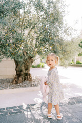 Naklejka premium Little girl with a toy rabbit stands in the yard near an olive tree, looking around. Back view
