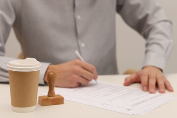 Obraz premium Man signing licensing agreement document at white table, focus on paper cup of drink and stamp
