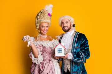Two elegant individuals in royal costumes showcasing a house model and keys against a vibrant...