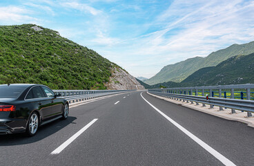 A sleek black sedan drives along a well-maintained highway winding through lush green mountains...