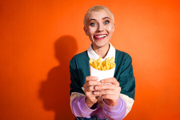 Energetic young woman in colorful attire holding a container of fries against a vibrant orange...