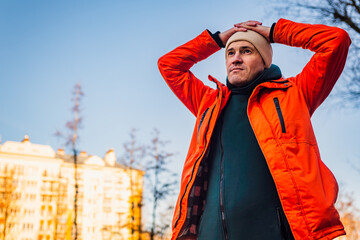Man in Bright Orange Jacket Standing Outdoors With Hands on Head Against Clear Sky