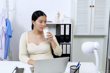 Asian pregnant woman in an office environment drinking a glass of milk, maintaining a healthy diet. Wearing a beige sweater and sitting at a desk with a laptop, balancing work and wellness.