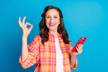 Cheerful senior woman showing OK gesture while holding smartphone, depicting happiness and modern...