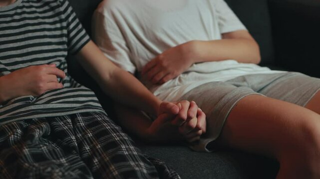 Two young people sit on a sofa, wearing casual home clothes, offering each other comfort and support through the simple act of holding hands